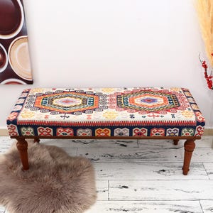 May include: A rectangular ottoman with a patterned fabric top in shades of red, blue, orange, and yellow. The wooden legs are dark brown. A decorative piece for a living room or bedroom.