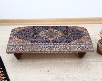May include: A rectangular footstool with a detailed, patterned rug top in shades of blue, cream, and red. The stool has dark wooden legs and a curved front edge. The rug design features floral and geometric motifs.