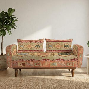 Może przedstawiać: Dwuosobowa sofa z tkaniny o geometrycznym wzorze w odcieniach terakoty, szałwiowej zieleni i kremu. Sofa posiada zaokrąglone podłokietniki, prostokątne oparcie z dwiema poduszkami oraz cztery nogi wykonane z ciemnobrązowego drewna.