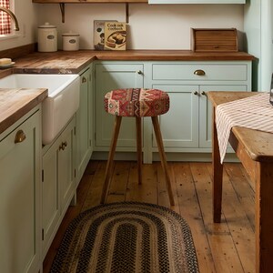 May include: A round stool with three wooden legs and a padded seat featuring a red, brown, and cream geometric pattern. The stool is positioned in a kitchen with light green cabinets, a wooden countertop, and a dark oval braided rug on the floor.
