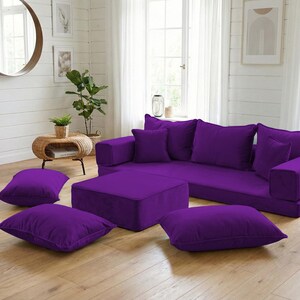 May include: A modular purple sectional sofa with matching throw pillows and an ottoman. The living room setting includes a round mirror, plants, and a woven basket, creating a modern and comfortable space.