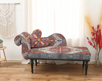 May include: A chaise lounge with a patterned fabric in shades of red, blue, and cream. The chaise has a curved back and armrest, and sits on black legs. A macrame wall hanging and a small side table are in the background.