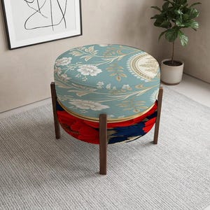 May include: A round ottoman featuring a light blue top cushion with a white and gold floral pattern, supported by a dark brown wooden frame. The lower section of the ottoman is upholstered in a vibrant fabric with red and dark blue floral motifs.