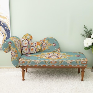 Puede incluir: Una chaise longue turquesa con un diseño estampado en naranja, amarillo y morado. La chaise longue tiene reposabrazos curvos y un cojín a juego. Las patas de madera son de color marrón oscuro. La chaise longue está sobre una alfombra blanca.