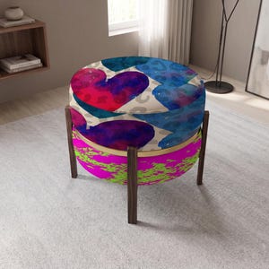 Könnte beinhalten: Ein runder Hocker mit einem Sitzkissen, das ein blau-rotes Herzmuster auf beigem Leopardenmuster zeigt. Der untere Teil ist mit einem leuchtend neonpinken und lindgrünen Textilstoff bezogen und steht auf vier dunkelbraunen Holzbeinen.