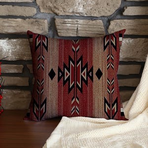 May include: Square cushion featuring a Southwestern geometric design in red, black, and beige. It rests on a wooden surface, against a stone wall, with a cream-coloured blanket draped nearby.