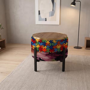 May include: A round ottoman with a wooden top and a gold metal rim. The upper section features a colorful puzzle piece pattern in red, blue, yellow, and green. The lower section displays a dark floral print. It stands on four black legs.