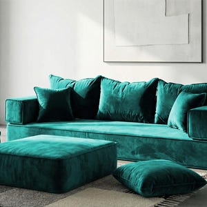 May include: A teal velvet sectional sofa with matching ottoman and throw pillows. The modern living room furniture set includes a large sofa, a square ottoman, and several square throw pillows in a rich teal color.