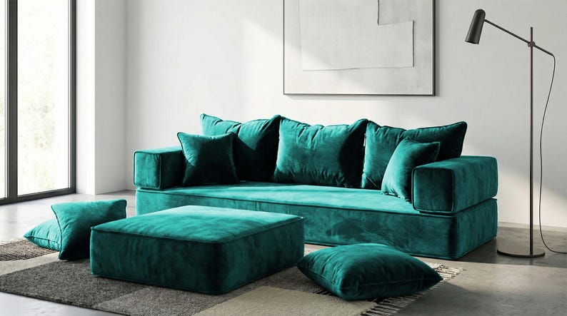 May include: A teal velvet sectional sofa with matching ottoman and throw pillows. The modern living room furniture set includes a large sofa, a square ottoman, and several square throw pillows in a rich teal color.