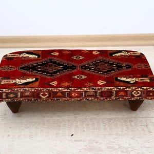 May include: A rectangular footstool with a red patterned top and dark wooden legs. The top features a central black diamond design and repeating patterns in shades of red, brown, and cream. The footstool is approximately 24 inches long.