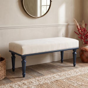May include: A cream-coloured upholstered bench with navy blue legs. The bench is in a room with a round mirror, a vase of red berries, and woven baskets. The room has a neutral colour palette.