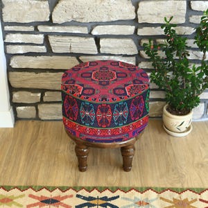 Può includere: Un pouf rotondo con un piano in tessuto fantasia nei toni del rosso, blu e verde. Il pouf ha gambe in legno scuro e poggia su un pavimento in legno chiaro. A destra c'è una piccola pianta in vaso. Il diametro è di circa 40 cm.