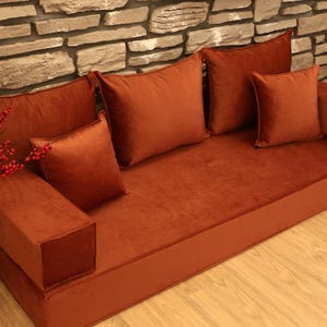 May include: A low-profile, rust-colored sofa with matching square throw pillows. The sofa has a simple, modern design with a plush, velvety texture. Decorative branches with red berries and tall, dried grass are in a vase to the left.