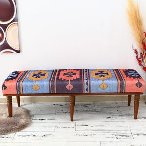Peut inclure: Un banc en bois avec un coussin à motifs colorés. Le coussin présente des motifs géométriques dans des tons de bleu, rouge, orange et jaune. Le banc a quatre pieds en bois et est posé sur un sol en bois blanc.