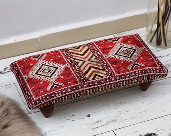 May include: Red and brown patterned footstool with wooden legs. The footstool has a geometric design with a red background and brown accents.