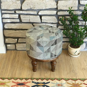 May include: A round upholstered ottoman with a geometric pattern in shades of gray, blue, and beige. The ottoman has a dark wooden base and four short, turned legs. A small potted plant sits nearby.