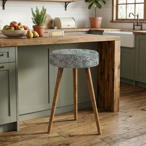 May include: A round stool featuring a blue upholstered seat with a white floral line-art pattern. The stool is supported by three slender, light-toned wooden legs that splay outward, providing a stable base for the circular, cushioned seat top.