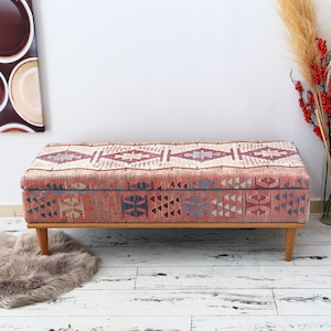 May include: A rectangular storage ottoman with a patterned fabric top in shades of red, blue, and cream. The ottoman has wooden legs and is placed on a light-colored wooden floor. Decorative elements include a faux fur rug and a vase of red berries.