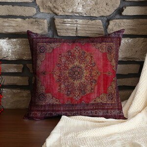 Könnte beinhalten: Ein quadratisches Zierkissen mit einem roten und bordeauxfarbenen orientalischen Teppichdesign. Das Kissen hat ein detailliertes Blumenmuster in der Mitte und einen Rand. Das Kissen liegt auf einer Holzoberfläche mit einem Hintergrund aus Steinziegeln.