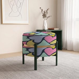 May include: A round ottoman featuring a dark blue fabric upholstery with a repeating pattern of hearts in shades of pink, yellow, and light green. The seat is supported by a dark frame with four legs and a thin gold-toned metal band accent.