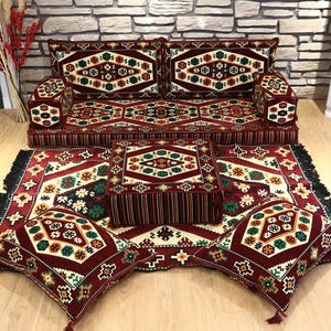 Op de afbeelding: Een complete vloerzitting set met een bank, twee armleuningen, een vierkante poef, twee vierkante kussens en een vloerkleed. De set heeft een bordeauxrood, cr&egrave;me en groen geometrisch patroon. Het vloerkleed heeft een zwarte franje.