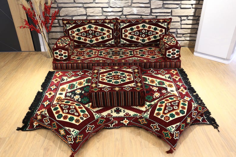 Op de afbeelding: Een complete vloerzitting set met een bank, twee armleuningen, een vierkante poef, twee vierkante kussens en een vloerkleed. De set heeft een bordeauxrood, cr&egrave;me en groen geometrisch patroon. Het vloerkleed heeft een zwarte franje.