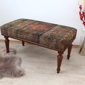May include: A rectangular ottoman with a patterned fabric top in shades of red, green, and brown. The ottoman has four dark wooden legs. A fluffy gray rug is on the floor in front of the ottoman.