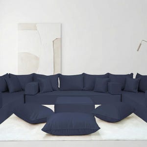 May include: A large, modular navy blue sectional sofa with multiple cushions and ottomans. The sofa is arranged in a U-shape on a white rug, with a square coffee table in the center. A potted plant is on the right side.
