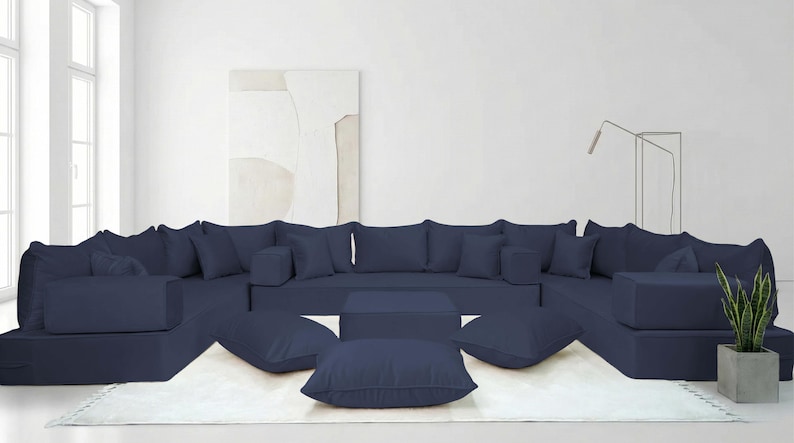 May include: A large, modular navy blue sectional sofa with multiple cushions and ottomans. The sofa is arranged in a U-shape on a white rug, with a square coffee table in the center. A potted plant is on the right side.