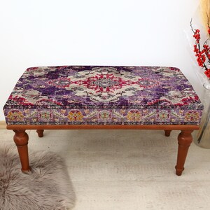 May include: A rectangular ottoman with a patterned top in shades of purple, red, and white. The wooden frame and legs are a warm brown color. The ottoman is a decorative furniture piece.