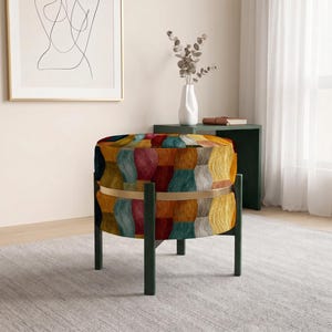 May include: A round ottoman featuring a multi-colored textured fabric with vertical brushstroke patterns in shades of orange, teal, red, and cream. The seat is encircled by a thin gold-toned metal band and supported by four dark green legs.