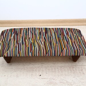 May include: A small footstool with a multi-coloured striped fabric top. The fabric has vertical stripes in shades of red, yellow, blue, and green. The stool has dark brown, angled wooden legs. The stool is approximately 60 cm long.