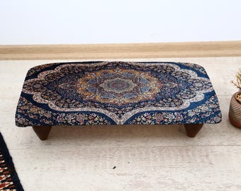 May include: A rectangular ottoman with a dark blue, ornate patterned top. The pattern features a central medallion with floral and geometric designs in shades of gold, blue, and cream. The ottoman rests on four short, dark wooden legs.