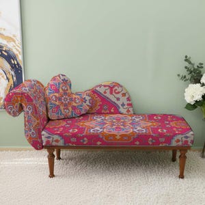 May include: A decorative chaise lounge with a vibrant, patterned fabric in shades of pink, orange, and blue. The wooden frame has four legs. The backrest and cushion feature a heart-shaped design. The room has a white rug and a painting.