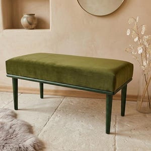 May include: A green velvet upholstered bench with a rectangular seat and four dark green legs. The bench is set against a neutral-toned wall with a decorative vase and dried plants.
