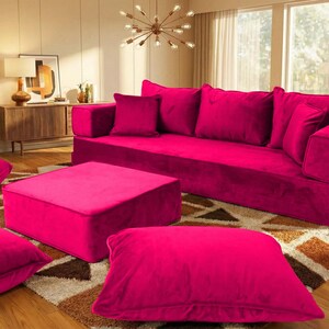 May include: A magenta velvet sofa with matching throw pillows and an ottoman. Several large floor cushions are scattered around the furniture. The room has a rug, plants, and a wooden cabinet.