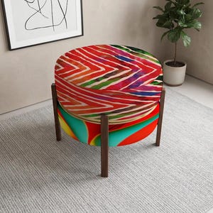 May include: A round ottoman featuring a top cushion with a vibrant red, white, and green chevron pattern. The base is upholstered in a colorful abstract design of red, yellow, and teal, supported by four dark brown wooden legs in a straight frame.