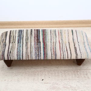 May include: A rectangular ottoman with a multi-colored striped pattern in shades of white, blue, red, and brown. The ottoman is supported by four dark brown wooden legs. A small potted plant sits nearby.
