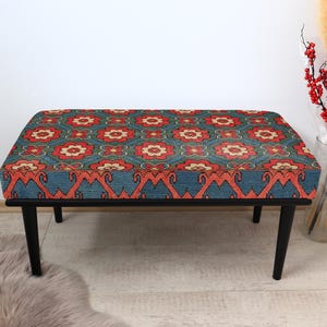 K&ouml;nnte beinhalten: Eine schwarze Holzbank mit einem gemusterten Kissen. Das Kissen hat ein rotes, oranges und blaues geometrisches Muster.