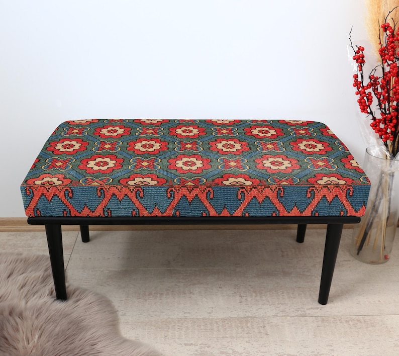 K&ouml;nnte beinhalten: Eine schwarze Holzbank mit einem gemusterten Kissen. Das Kissen hat ein rotes, oranges und blaues geometrisches Muster.