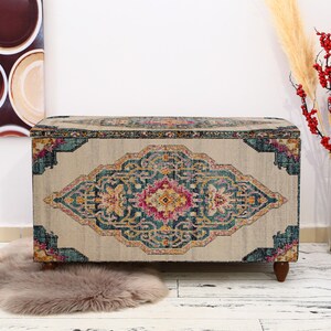 May include: A rectangular storage ottoman with a beige and patterned top. The pattern features a central medallion in pink, yellow, and teal. The ottoman has dark wooden legs and is placed on a white floor.