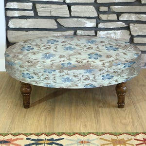 May include: An upholstered ottoman with a floral pattern in blue and brown on a cream background. The round ottoman has four dark wooden legs. The ottoman is placed on a light wood floor, with a patterned rug visible in the foreground.