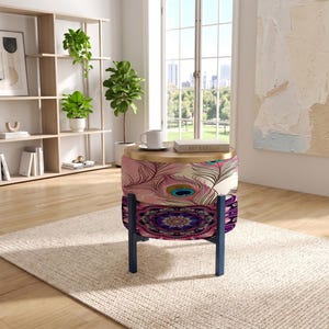 May include: A round ottoman with a wooden top featuring a peacock feather pattern on the upper section and a purple mandala design on the lower section, supported by four dark blue legs. A white mug and a book are placed on the wooden surface.