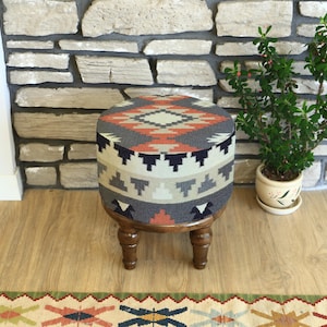 May include: A round upholstered ottoman with a geometric pattern in shades of gray, orange, and cream. The ottoman has dark wooden legs and sits on a wooden floor. A small potted plant is to the right.