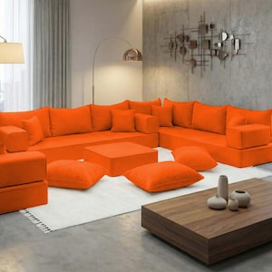 May include: An orange sectional sofa with matching square ottomans and throw pillows. The modern living room also features a wooden coffee table, a white rug, and a floor lamp. The room has large windows with sheer curtains.