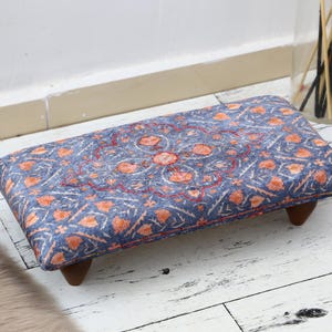 May include: A blue and orange patterned footstool with wooden legs. The footstool is sitting on a white wooden floor.