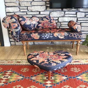 May include: A navy blue settee and heart-shaped ottoman with floral upholstery in orange, red, and gold. The settee has curved arms and wooden legs. The ottoman sits on a wooden base. A patterned rug is in the foreground.