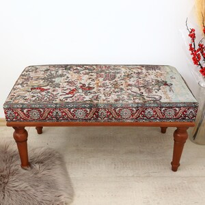 May include: A wooden ottoman with a patterned fabric top. The fabric has a floral design with red, blue, and green colours. The ottoman has four legs.