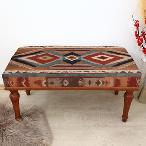 May include: A wooden ottoman with a colourful geometric patterned fabric top. The ottoman has four legs and is sitting on a light brown rug.