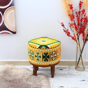 May include: A round, yellow ottoman with a geometric pattern in green, black, and white. The ottoman has a wooden base with four short, dark brown legs. The top is a bright yellow with a central green and white design.